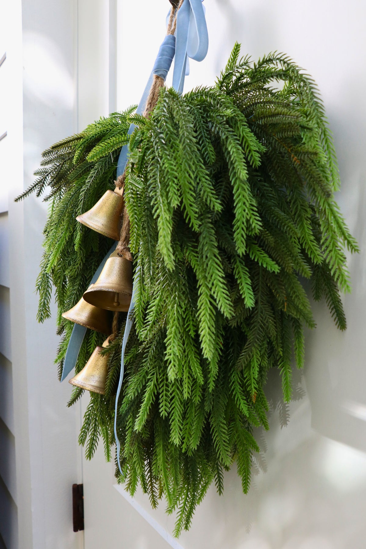 Cascading faux Norfolk pine wreath with dusty blue velvet ribbon and five gold-tone bells on rustic cord.
