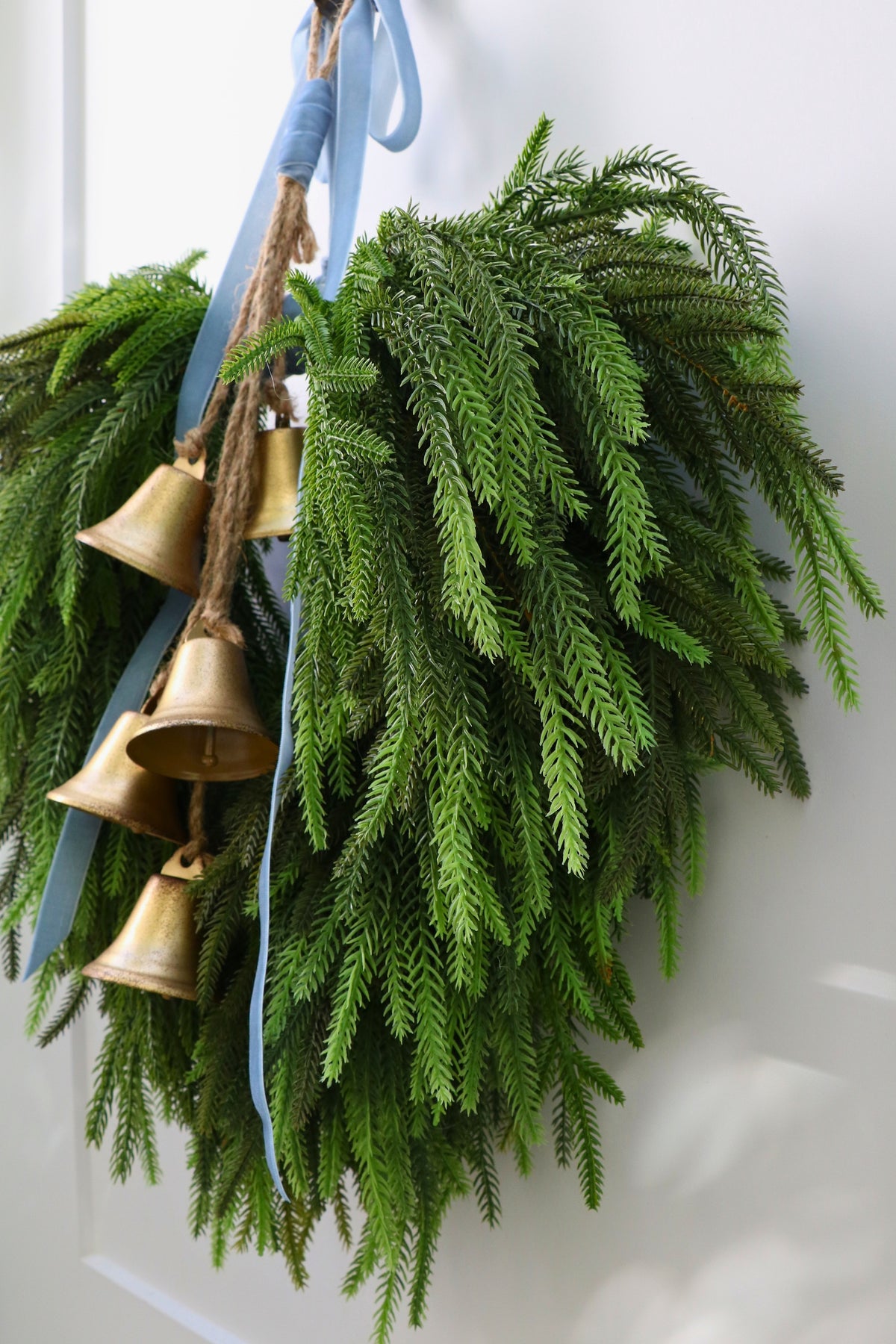 Cascading faux Norfolk pine wreath with dusty blue velvet ribbon and five gold-tone bells on rustic cord.