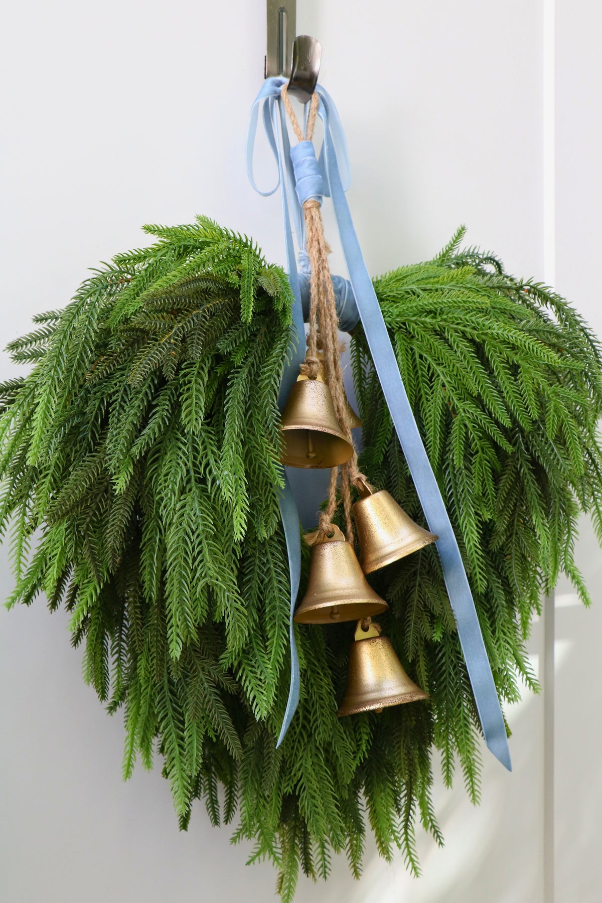 Cascading faux Norfolk pine wreath with dusty blue velvet ribbon and five gold-tone bells on rustic cord.