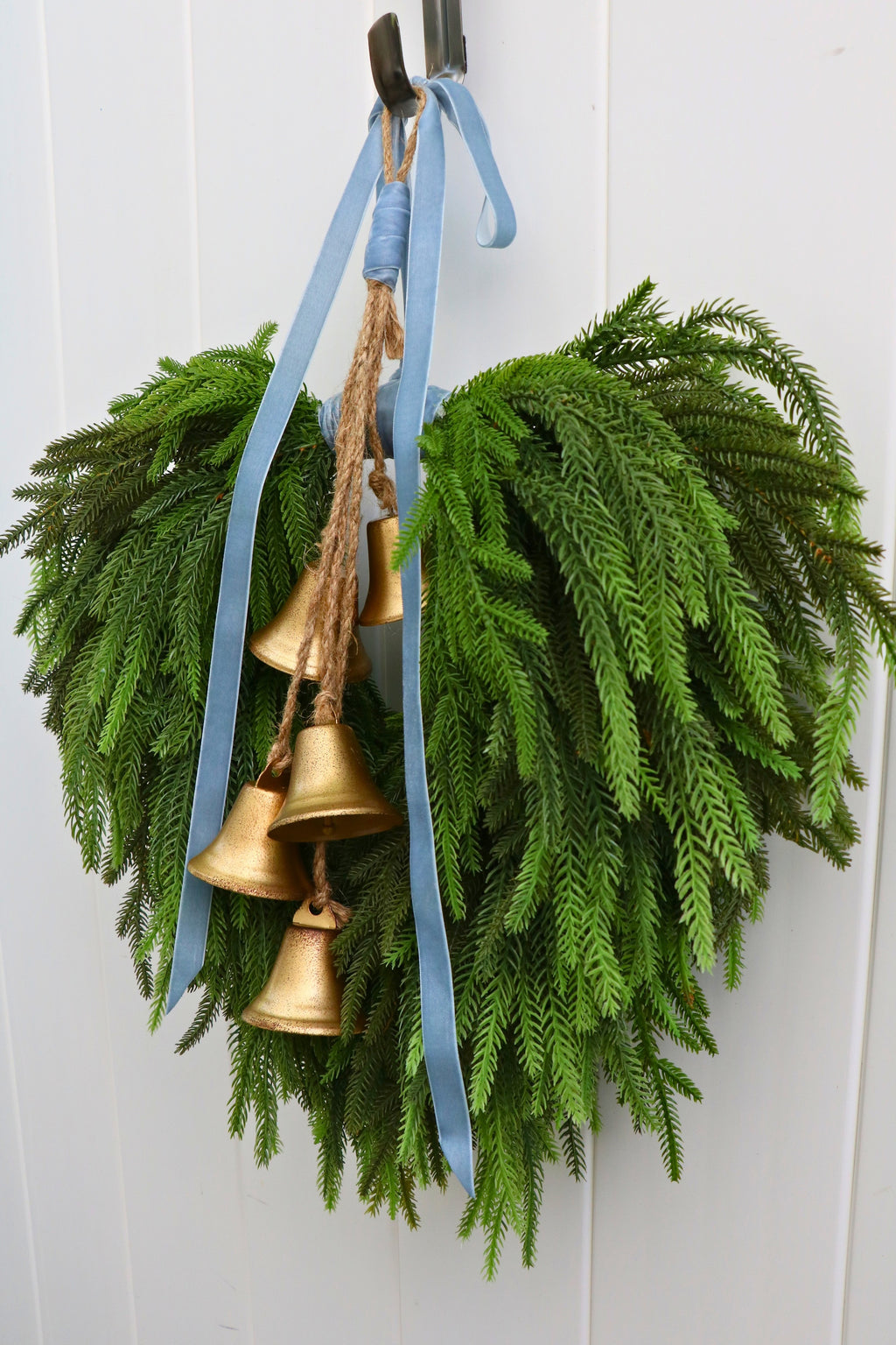 Cascading faux Norfolk pine wreath with dusty blue velvet ribbon and five gold-tone bells on rustic cord.