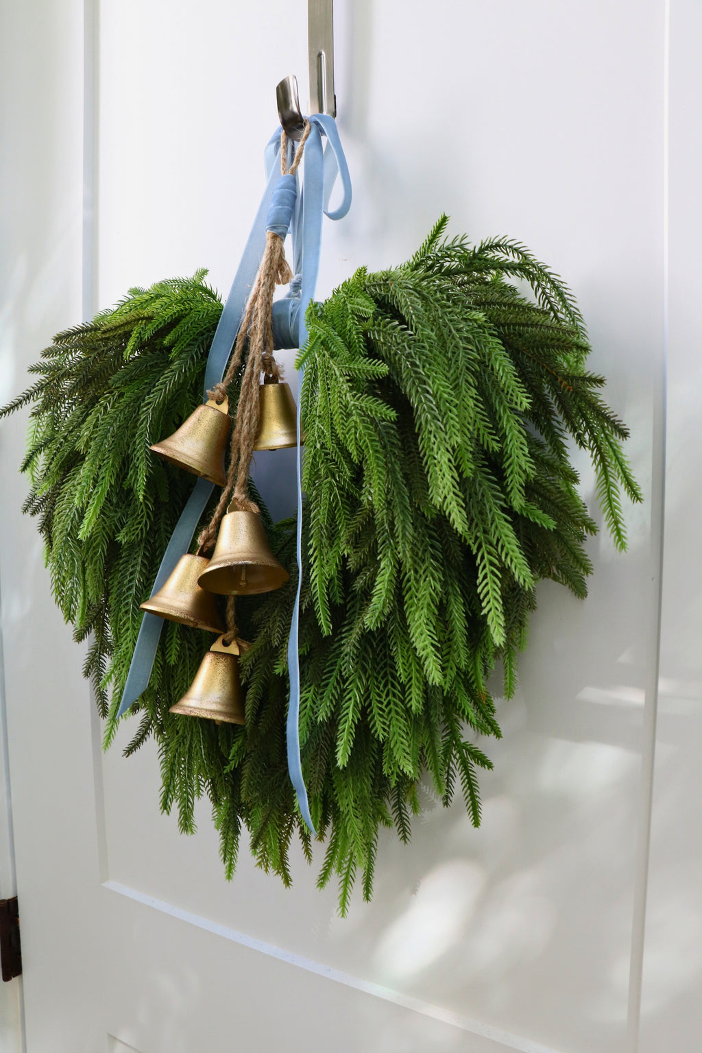 Cascading faux Norfolk pine wreath with dusty blue velvet ribbon and five gold-tone bells on rustic cord.