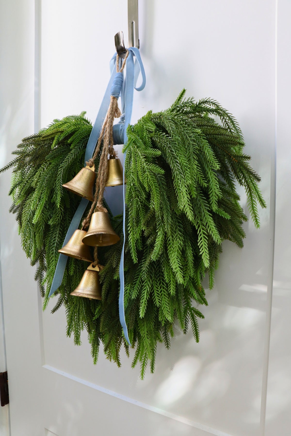 Cascading faux Norfolk pine wreath with dusty blue velvet ribbon and five gold-tone bells on rustic cord.