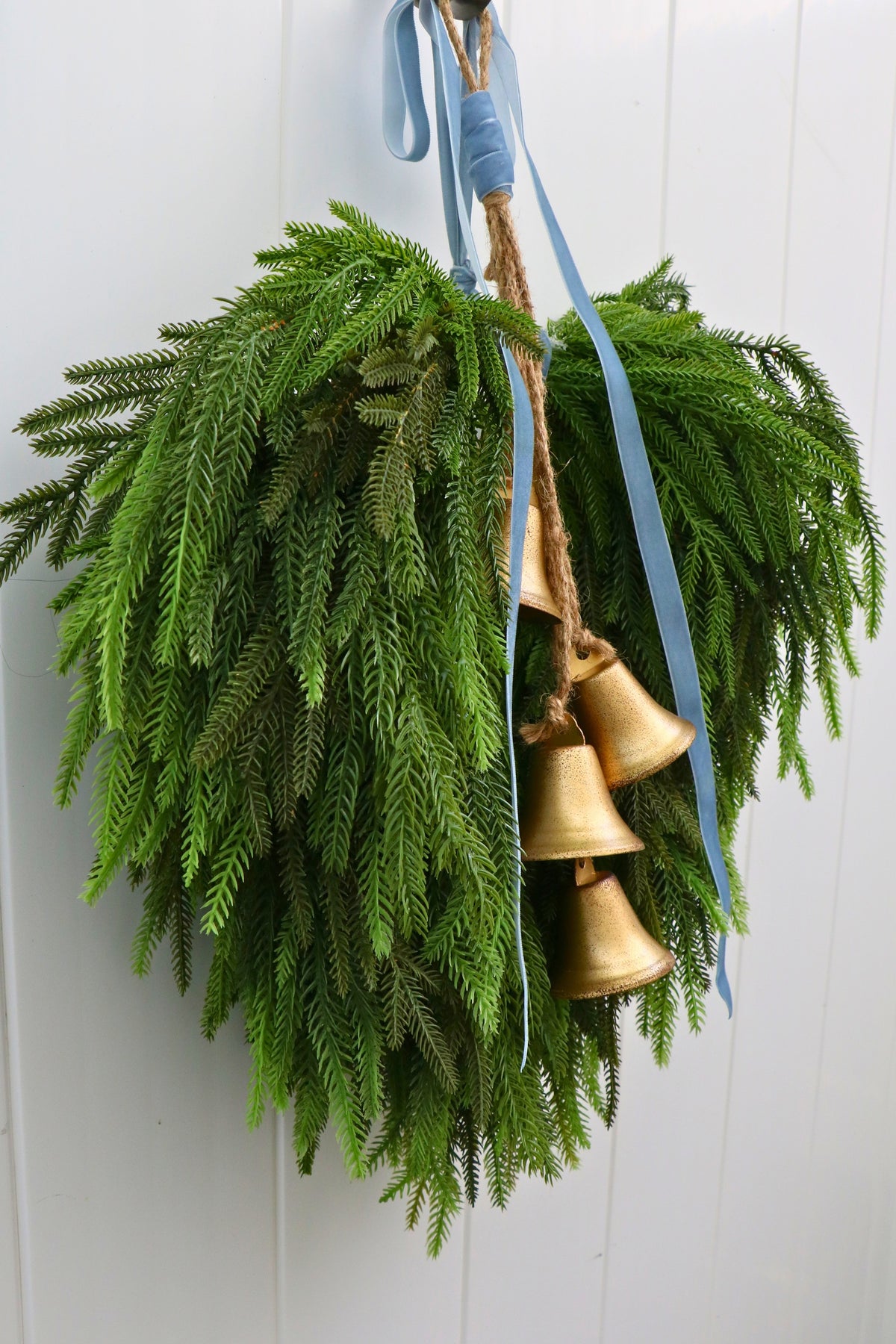 Cascading faux Norfolk pine wreath with dusty blue velvet ribbon and five gold-tone bells on rustic cord.