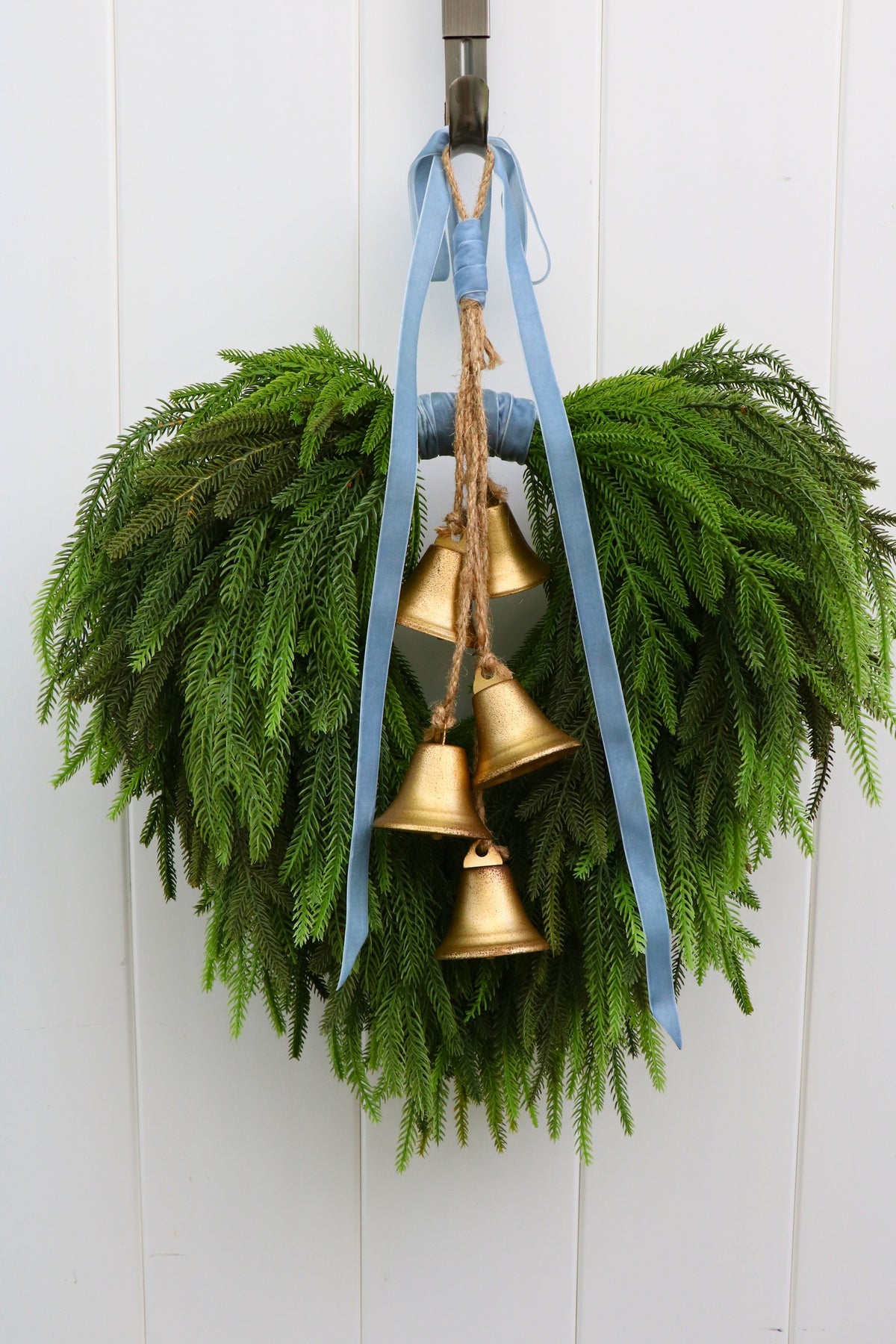 Cascading faux Norfolk pine wreath with dusty blue velvet ribbon and five gold-tone bells on rustic cord.