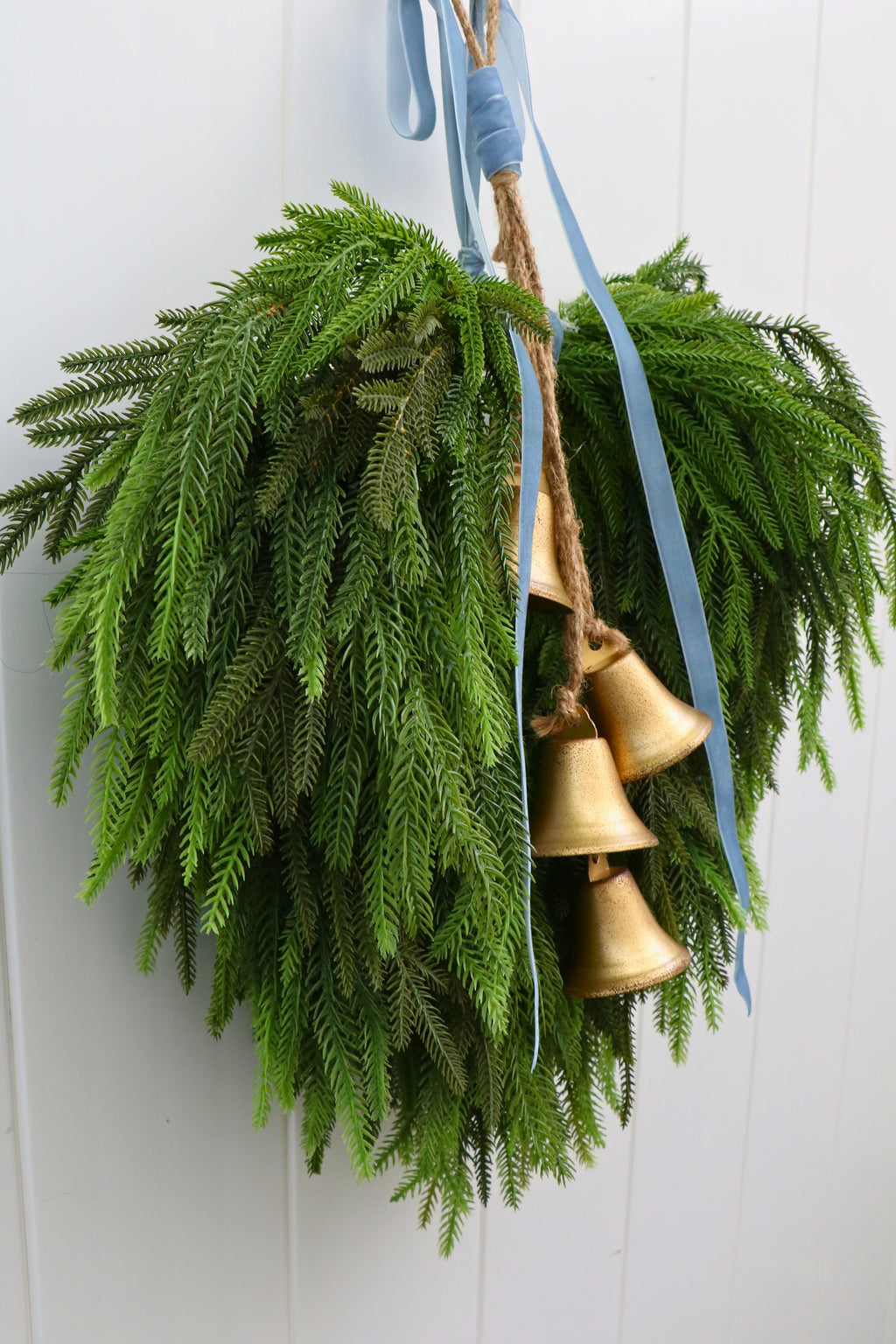 Cascading faux Norfolk pine wreath with dusty blue velvet ribbon and five gold-tone bells on rustic cord.
