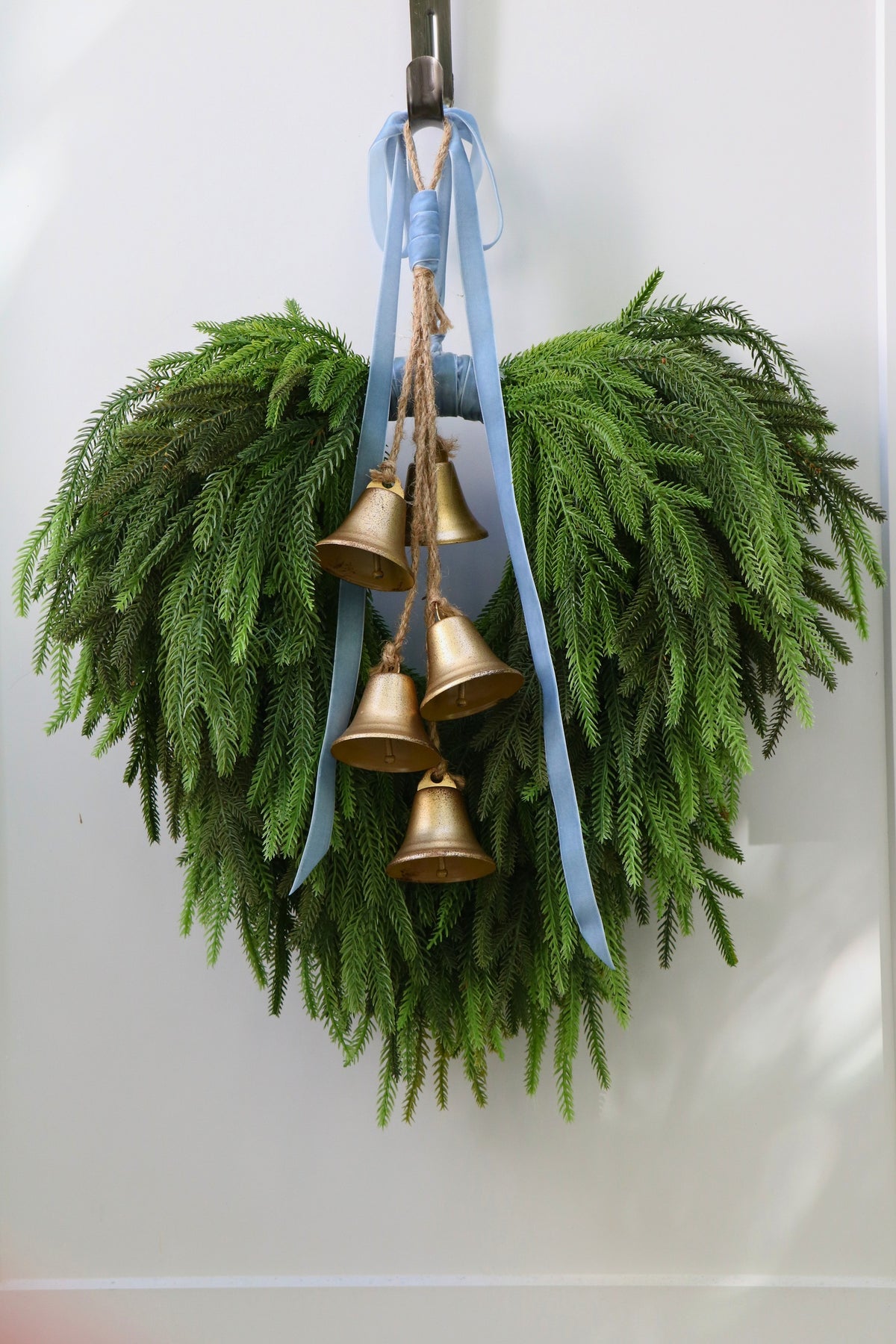 Cascading faux Norfolk pine wreath with dusty blue velvet ribbon and five gold-tone bells on rustic cord.