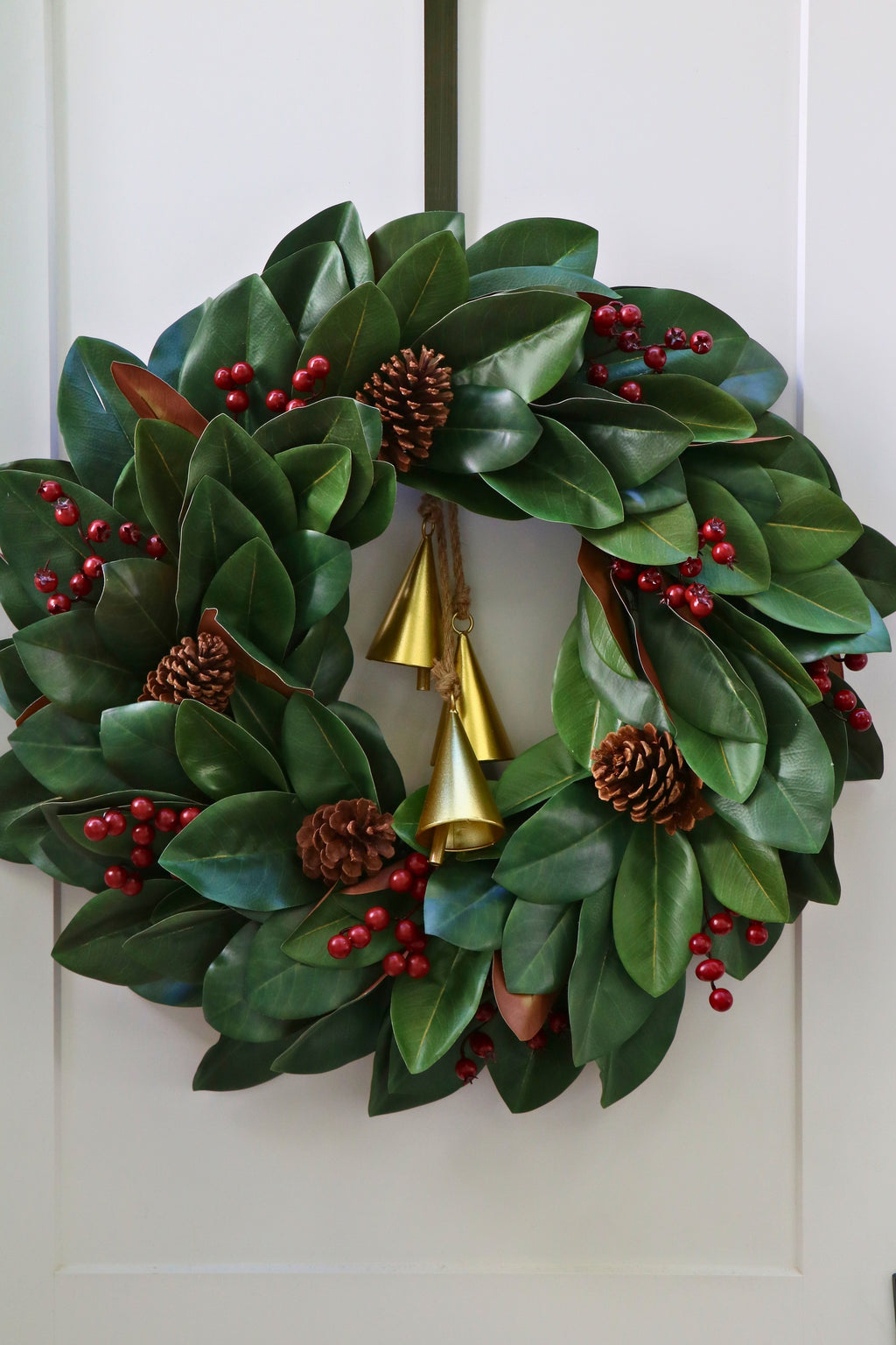 Magnolia leaf wreath with red berry clusters, four pinecones and three hanging gold-tone bells.