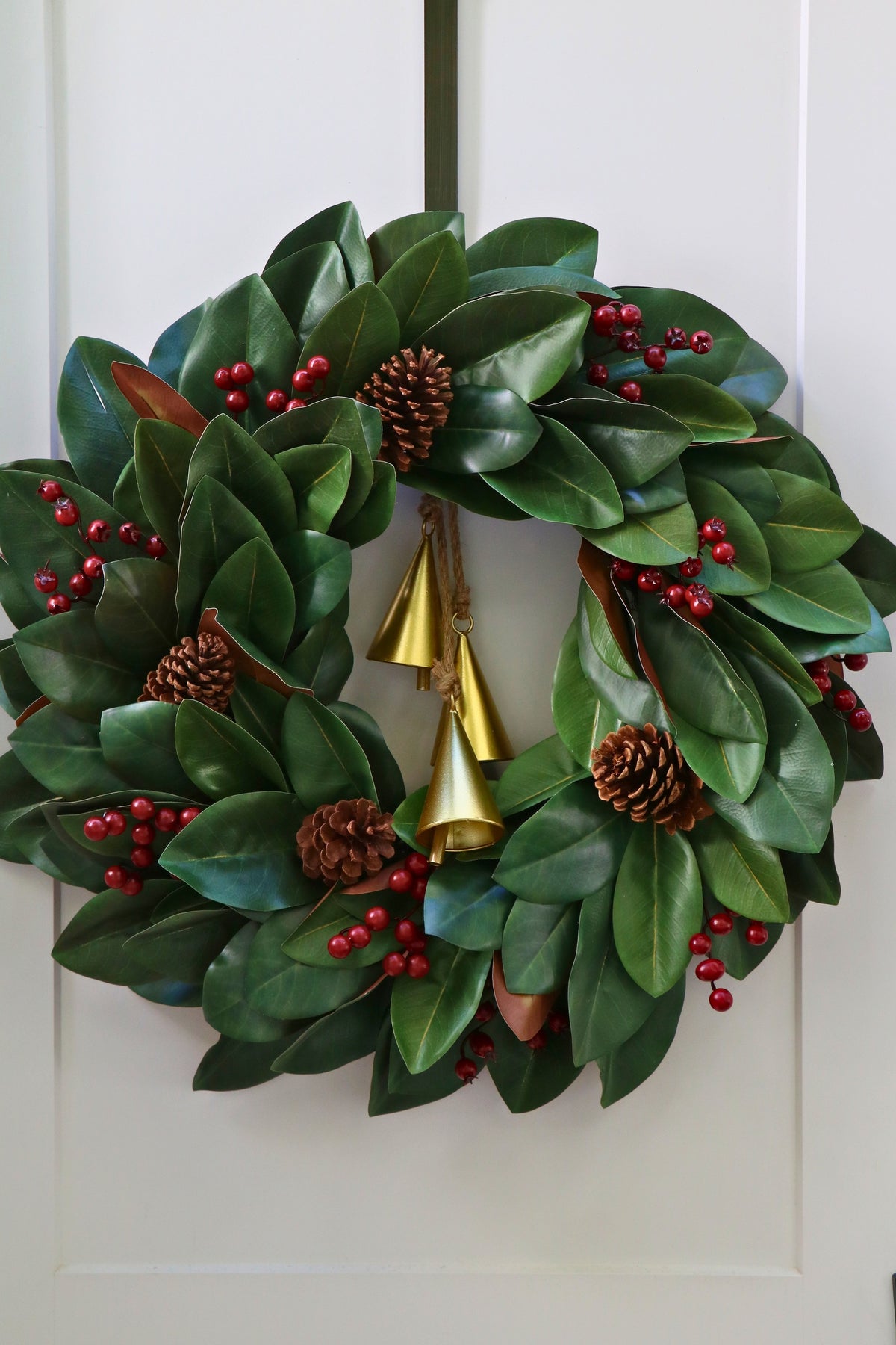 Magnolia leaf wreath with red berry clusters, four pinecones and three hanging gold-tone bells.