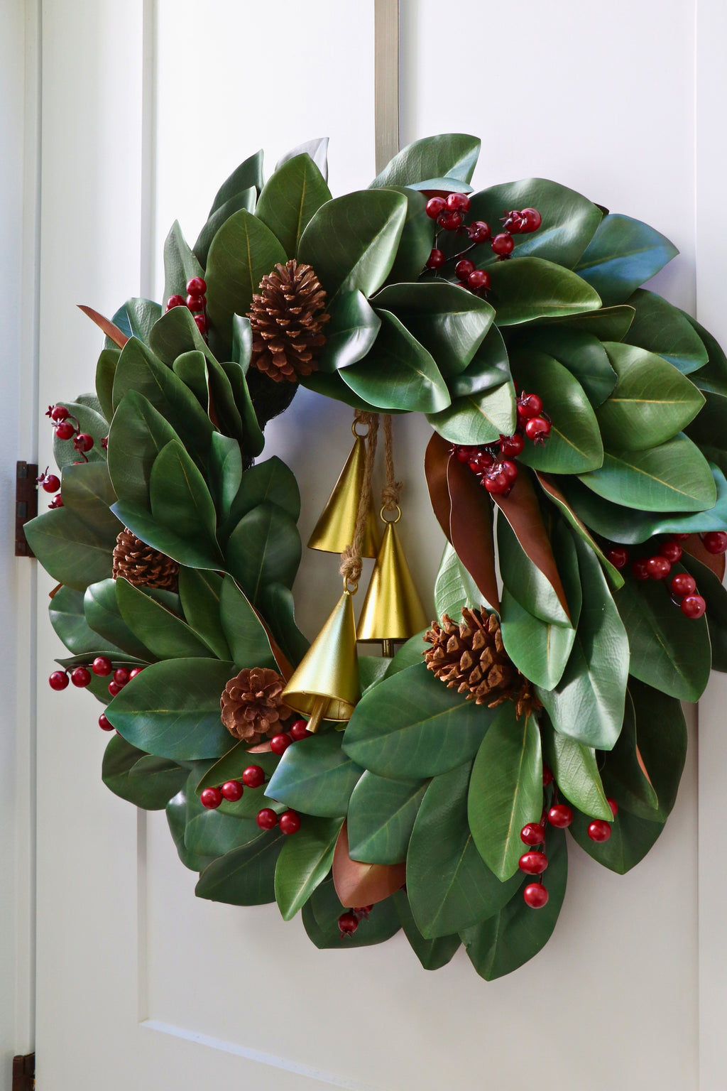 Magnolia leaf wreath with red berry clusters, four pinecones and three hanging gold-tone bells.