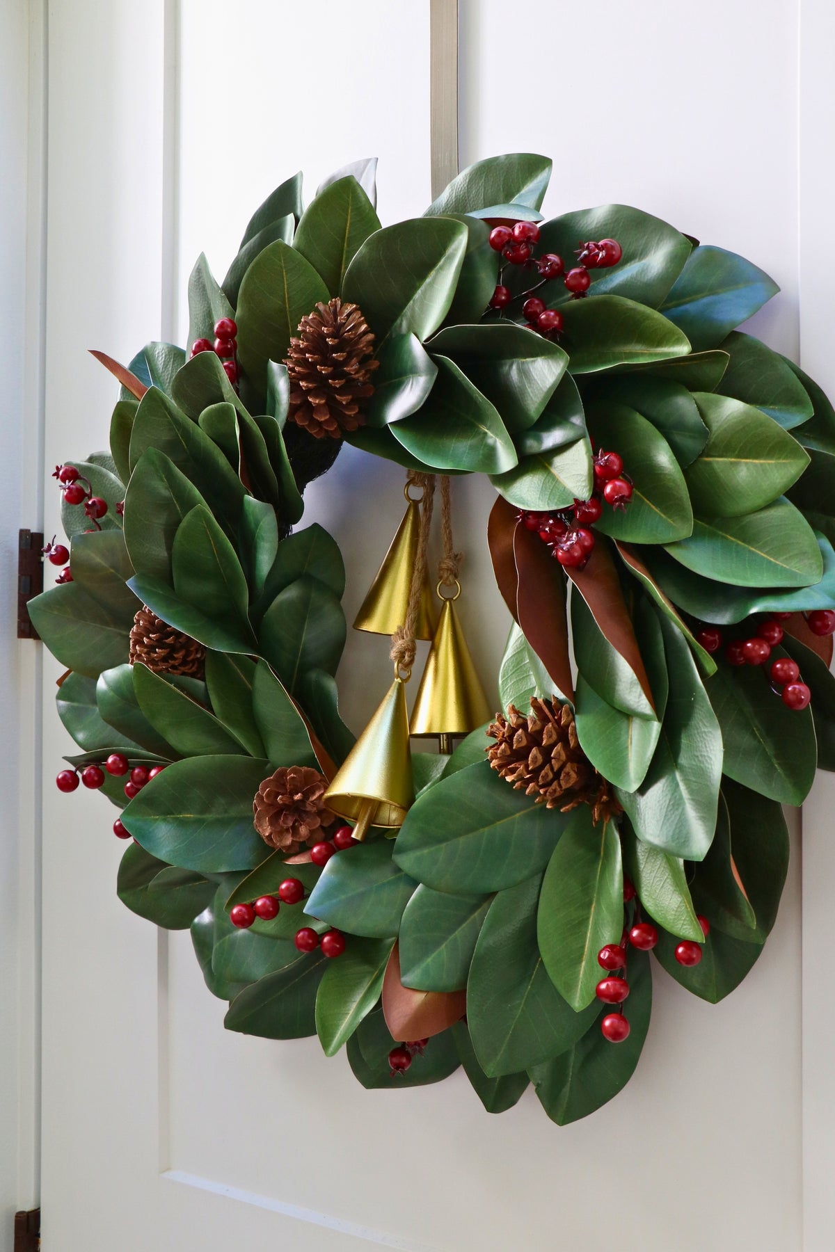 Magnolia leaf wreath with red berry clusters, four pinecones and three hanging gold-tone bells.