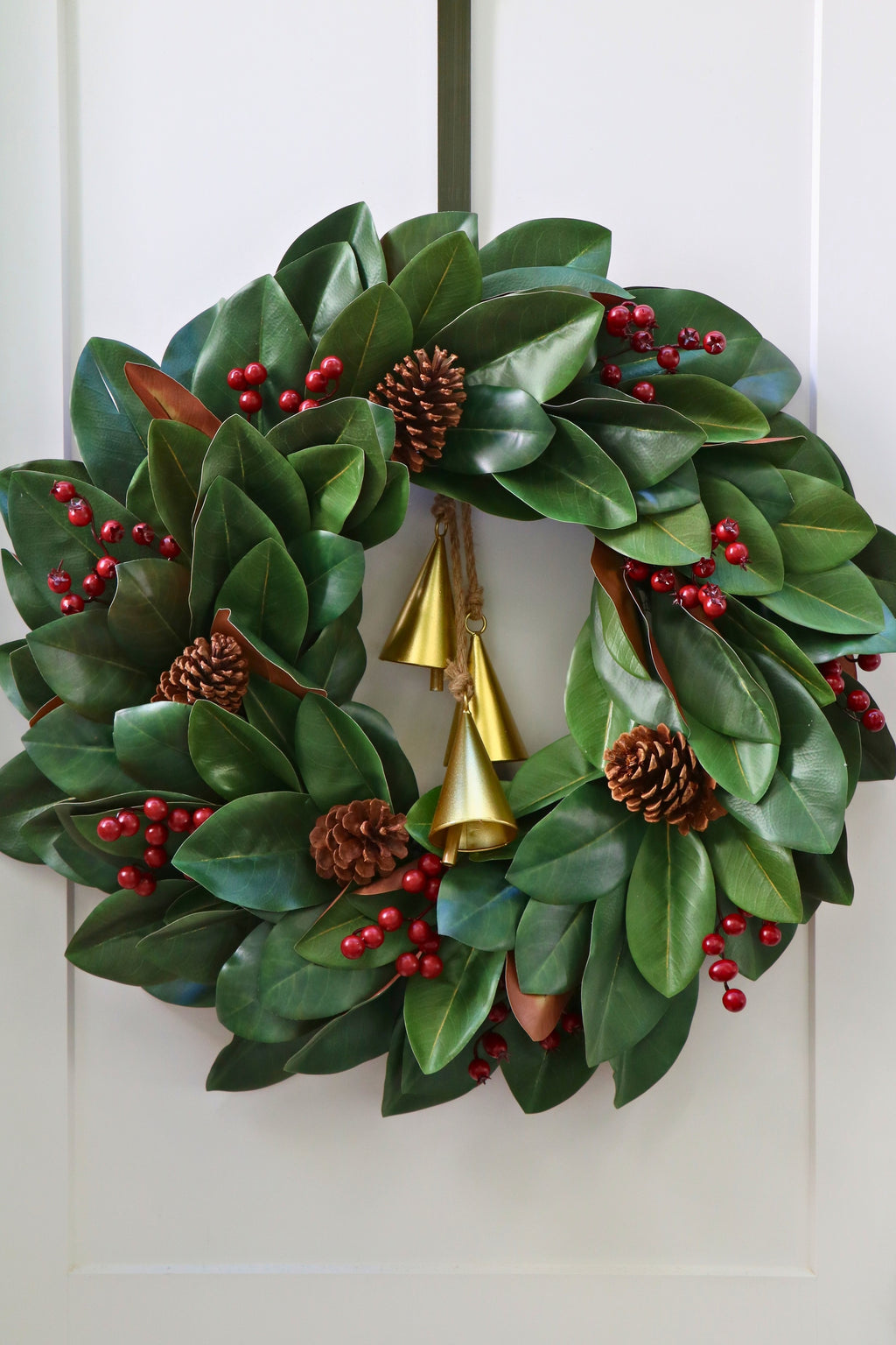 Magnolia leaf wreath with red berry clusters, four pinecones and three hanging gold-tone bells.