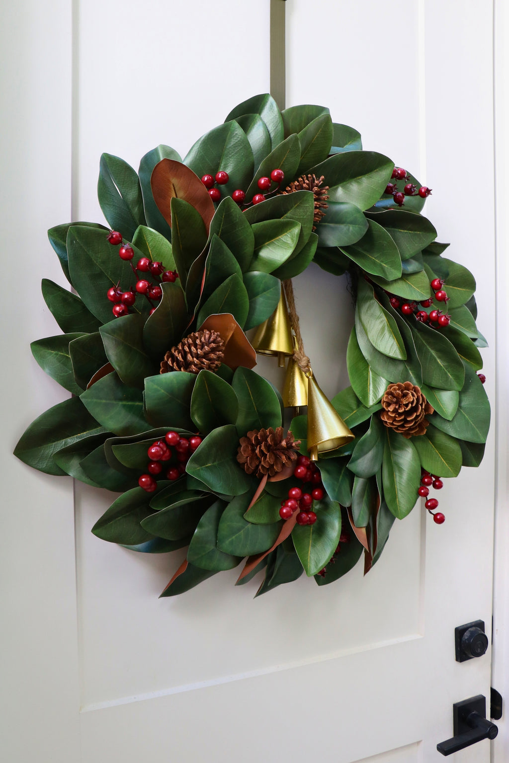 Magnolia leaf wreath with red berry clusters, four pinecones and three hanging gold-tone bells.