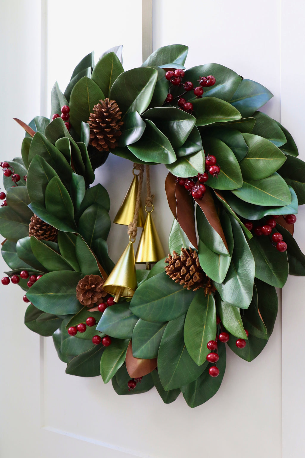 Magnolia leaf wreath with red berry clusters, four pinecones and three hanging gold-tone bells.