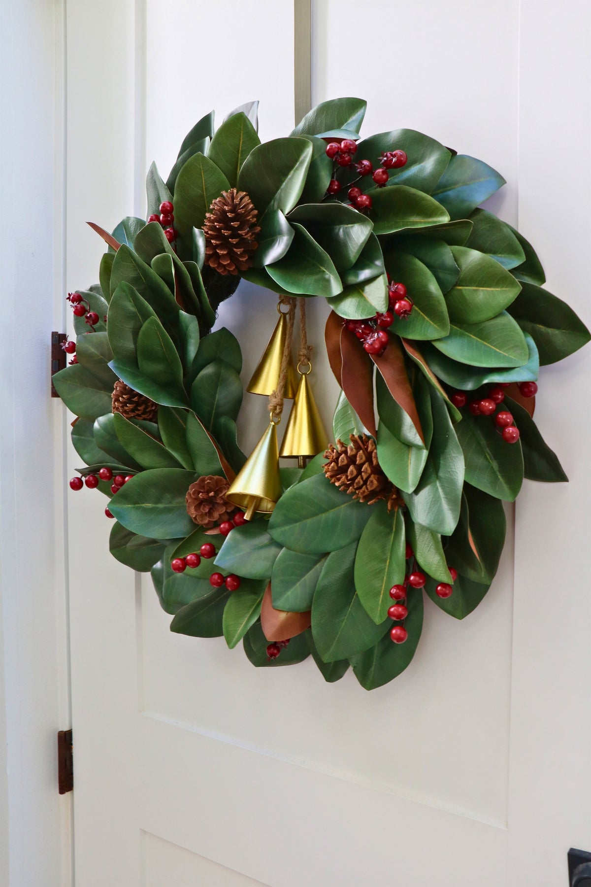 Magnolia leaf wreath with red berry clusters, four pinecones and three hanging gold-tone bells.