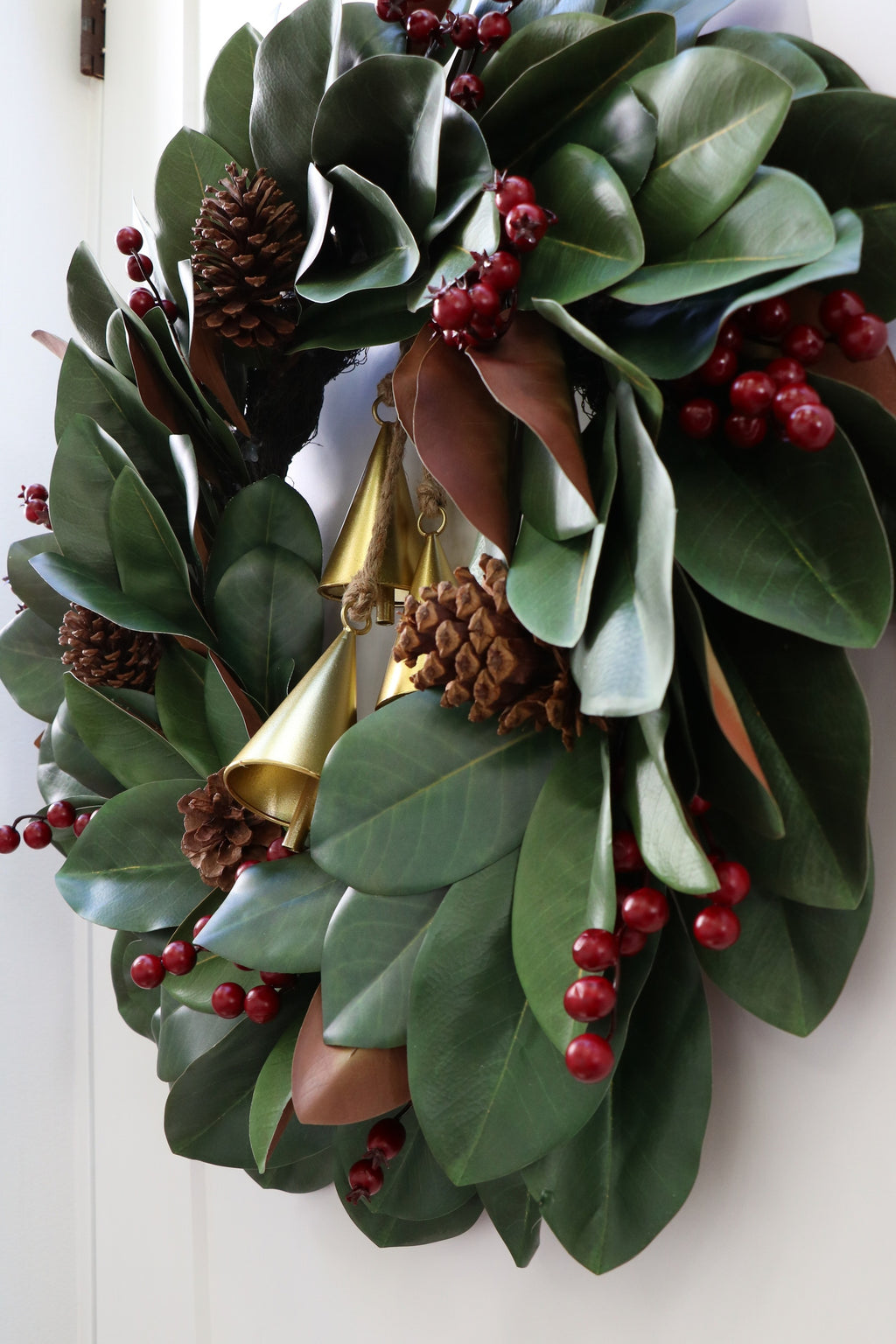 Magnolia leaf wreath with red berry clusters, four pinecones and three hanging gold-tone bells.