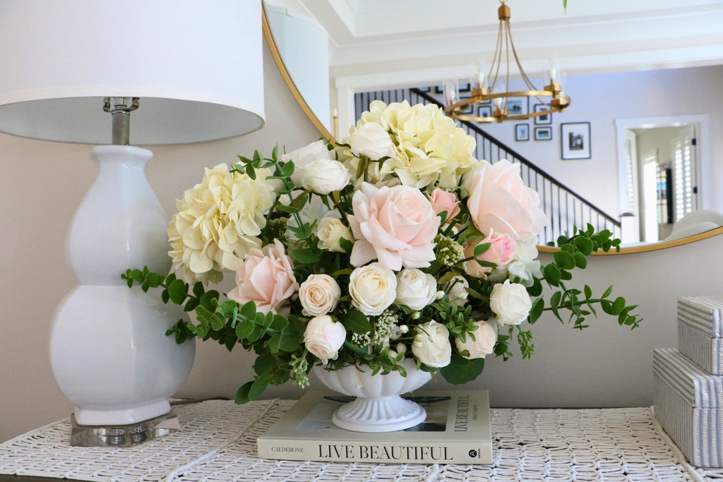 Ivory and blush rose and hydrangea arrangement with eucalyptus in a white pedestal-style compote vase.
