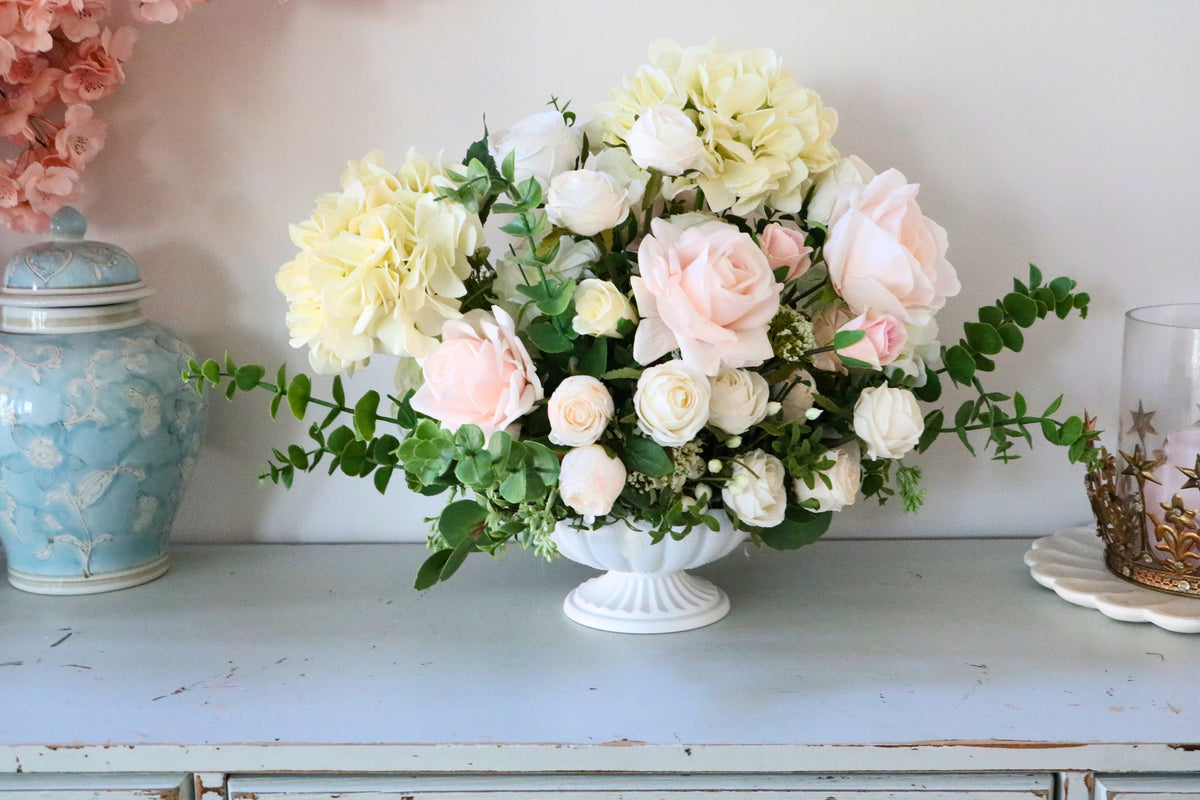 Ivory and blush rose and hydrangea arrangement with eucalyptus in a white pedestal-style compote vase.