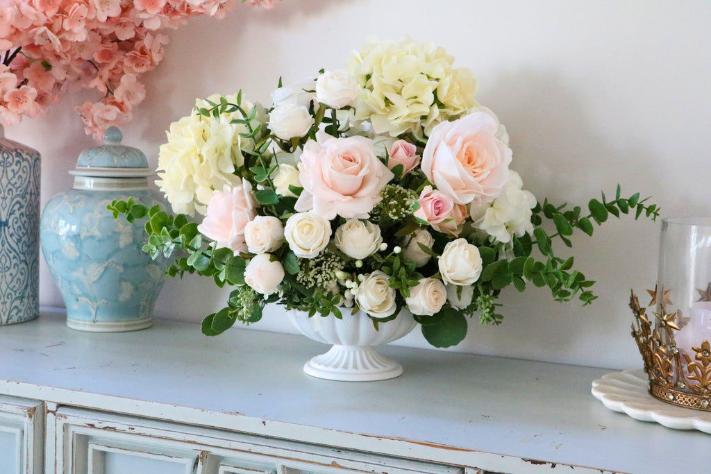 Ivory and blush rose and hydrangea arrangement with eucalyptus in a white pedestal-style compote vase.