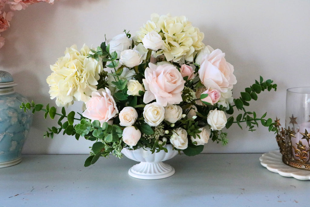 Ivory and blush rose and hydrangea arrangement with eucalyptus in a white pedestal-style compote vase.