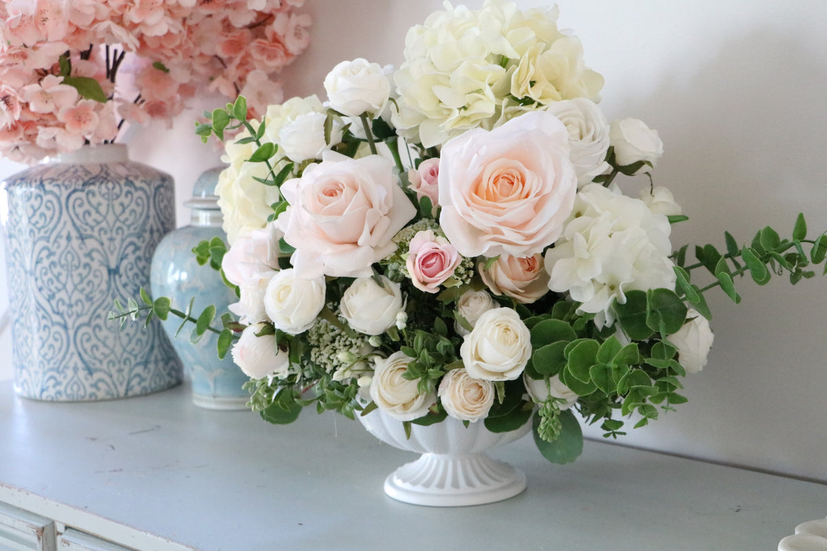 Ivory and blush rose and hydrangea arrangement with eucalyptus in a white pedestal-style compote vase.
