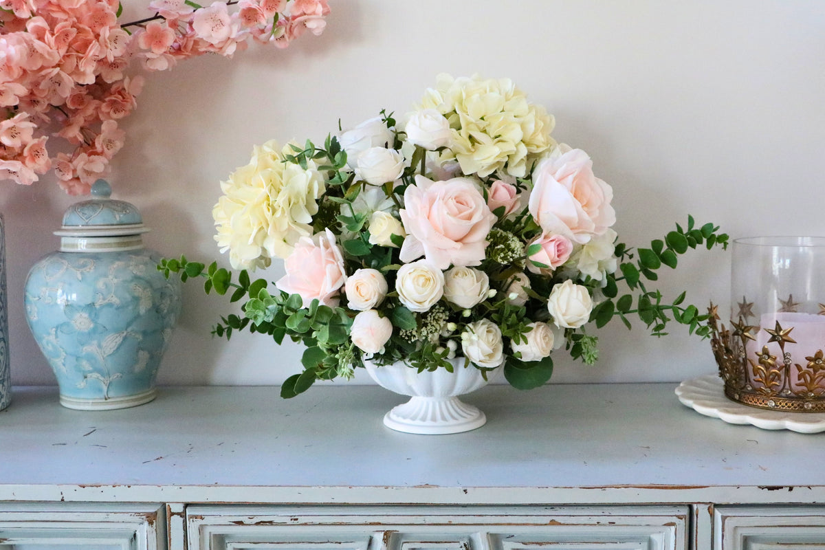 Ivory and blush rose and hydrangea arrangement with eucalyptus in a white pedestal-style compote vase.