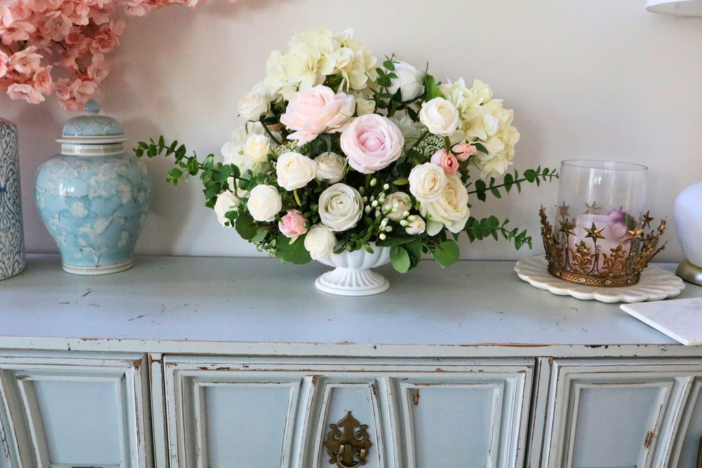 Ivory and blush rose and hydrangea arrangement with eucalyptus in a white pedestal-style compote vase.