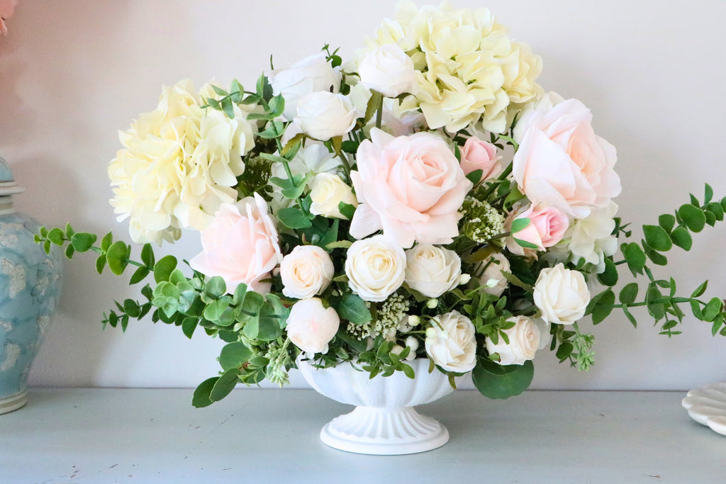 Ivory and blush rose and hydrangea arrangement with eucalyptus in a white pedestal-style compote vase.