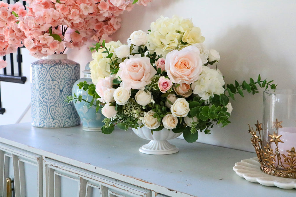 Ivory and blush rose and hydrangea arrangement with eucalyptus in a white pedestal-style compote vase.