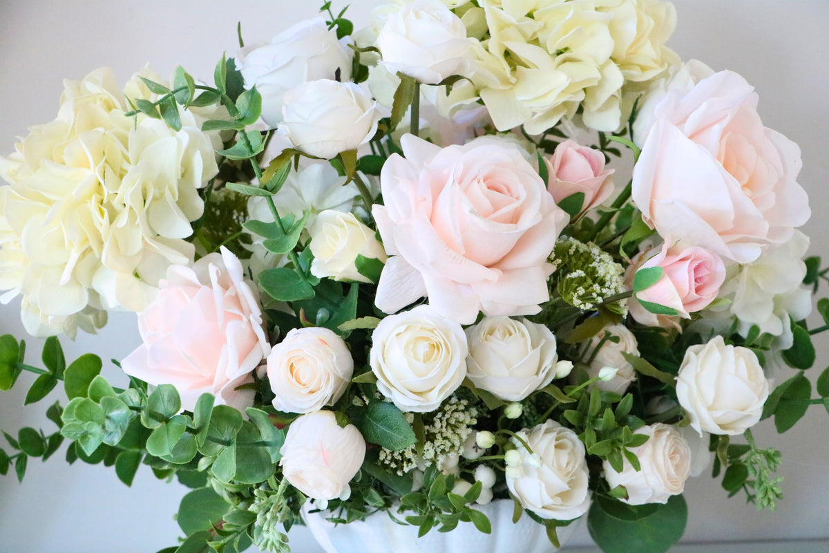 Ivory and blush rose and hydrangea arrangement with eucalyptus in a white pedestal-style compote vase.