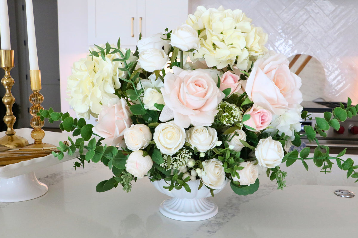 Ivory and blush rose and hydrangea arrangement with eucalyptus in a white pedestal-style compote vase.
