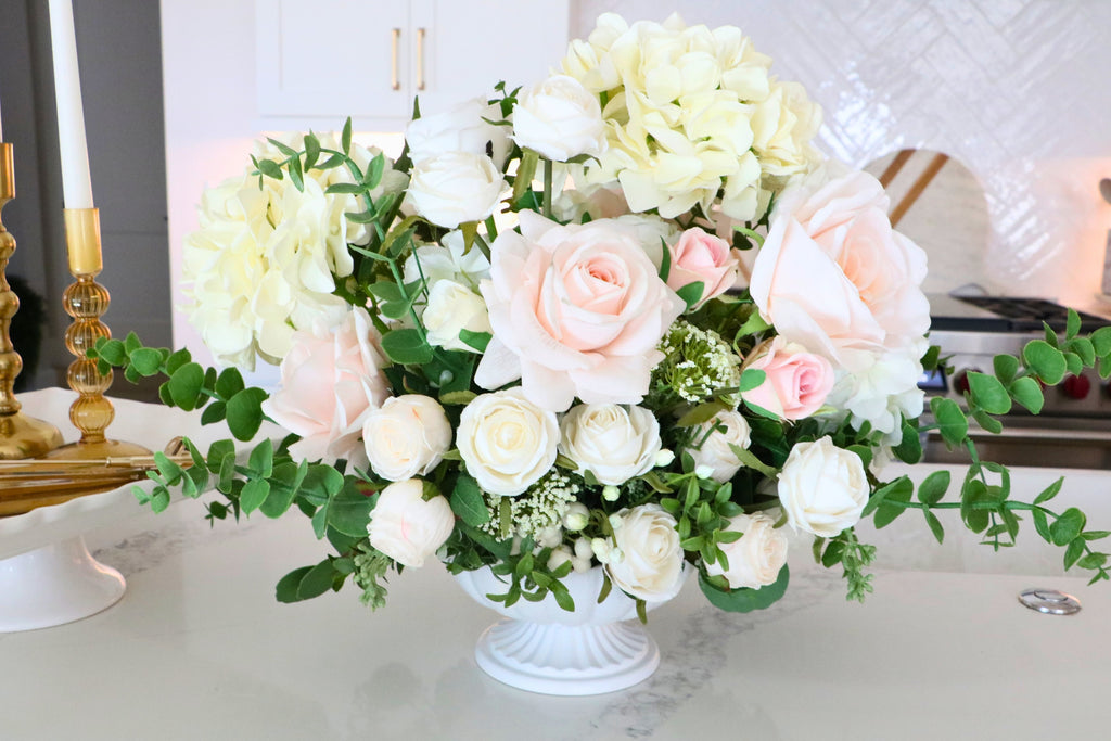 Ivory and blush rose and hydrangea arrangement with eucalyptus in a white pedestal-style compote vase.