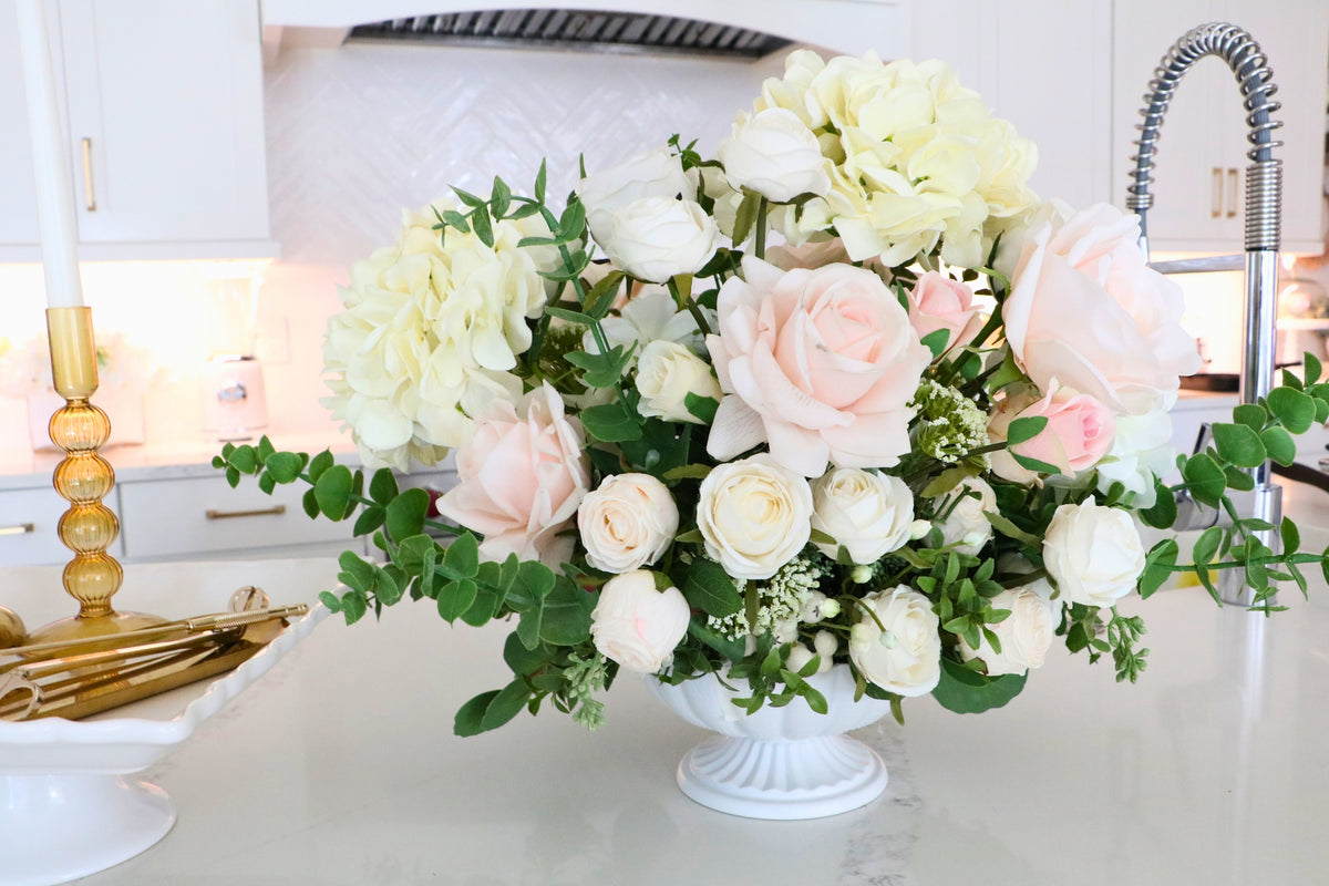 Ivory and blush rose and hydrangea arrangement with eucalyptus in a white pedestal-style compote vase.