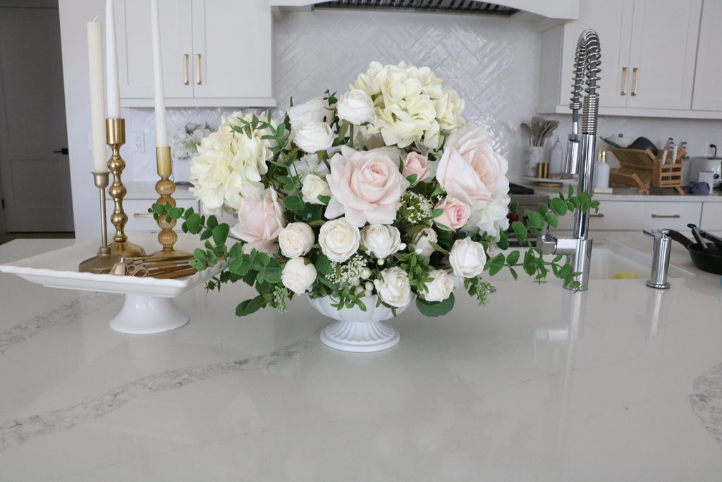 Ivory and blush rose and hydrangea arrangement with eucalyptus in a white pedestal-style compote vase.