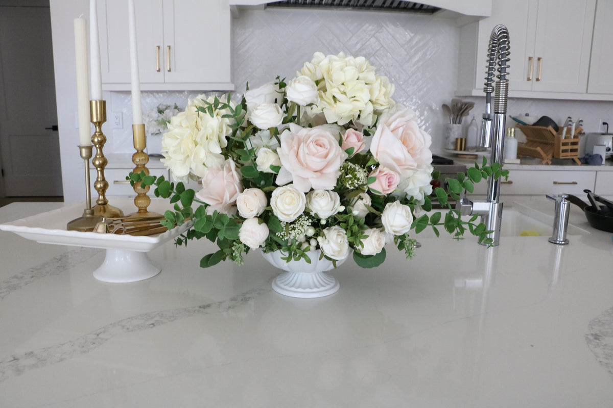Ivory and blush rose and hydrangea arrangement with eucalyptus in a white pedestal-style compote vase.
